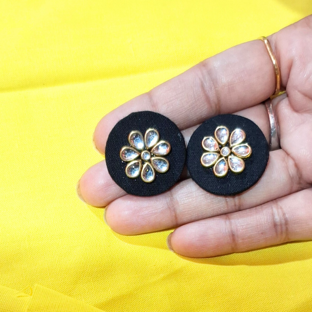 Hand holding round black fabric earrings with gold floral crystal design on yellow background