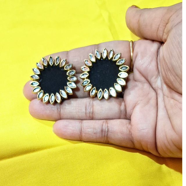 Hand holding round black stud earrings with gold and crystal petal border on yellow background