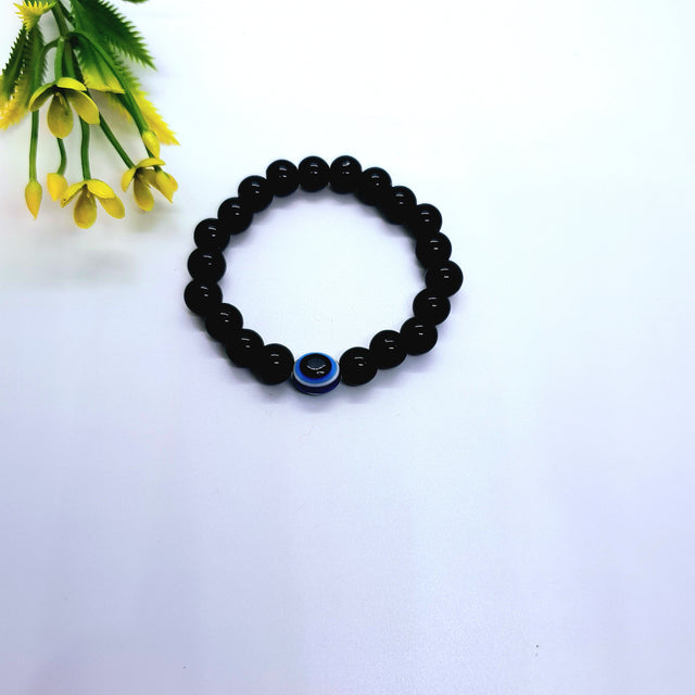 Black beaded bracelet with blue evil eye charm on white background, beside yellow flowers