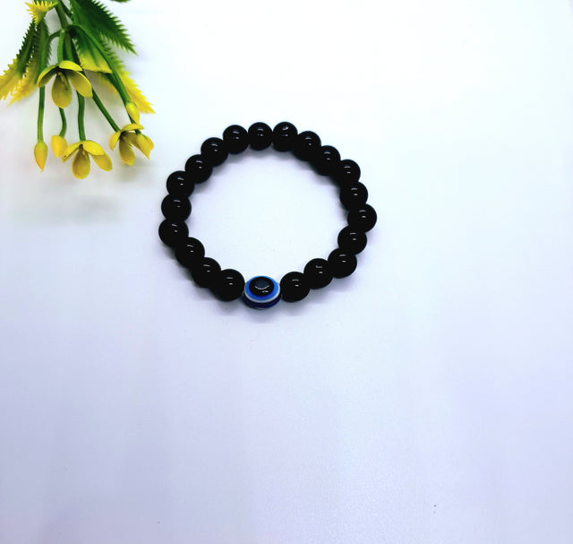 Black beaded bracelet with blue evil eye charm on white background, beside yellow flowers